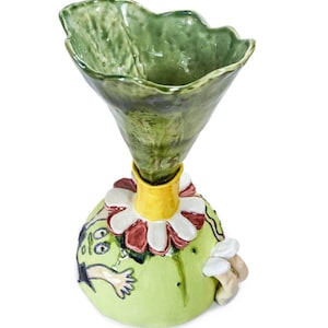 Puede incluir: Un jarrón de cerámica verde con un diseño único. El jarrón tiene forma de flor con una boca ancha y abierta y una base estrecha. La base está decorada con una cara de dibujos animados caprichosa y un borde amarillo.