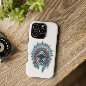 May include: White phone case featuring a Native American headdress design with blue feathers and an orange and blue emblem. The case is on a wooden surface, with a camera and plant in the background.