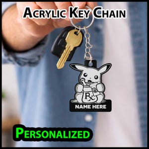 May include: An acrylic keychain featuring a black and white graphic of a cartoon creature holding a mortar and pestle with an Rx symbol. The keychain includes a metal ring, a gold-colored key, and the text "NAME HERE" printed on the bottom.