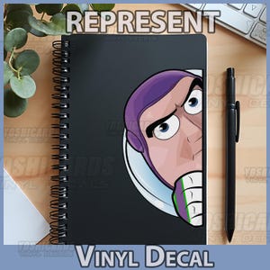 May include: A black spiral-bound notebook featuring a Buzz Lightyear graphic. The notebook is accompanied by a black pen. The word "REPRESENT" is at the top, and "VINYL DECAL" is at the bottom.