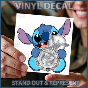May include: A blue vinyl decal featuring the character Stitch from Lilo & Stitch, holding a silver emblem with an eagle and the text "Security Police Department of the Air Force". The decal also includes the text "Vinyl Decal" and "Stand Out & Represent".