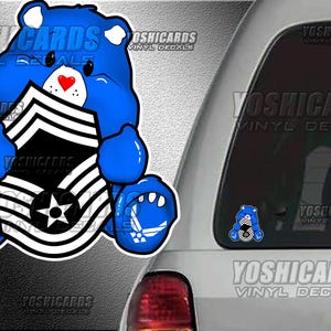 May include: A blue cartoon bear sticker holding a black and white emblem with a star. The bear has a red heart nose and Air Force logos on its paws. The sticker is on a car window.