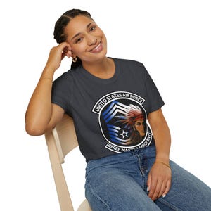 May include: Dark gray t-shirt featuring a circular emblem with the text "UNITED STATES AIR FORCE CHIEF MASTER SERGEANT." The emblem includes a Native American woman in a headdress and blue and white stripes. The shirt is worn by a person.