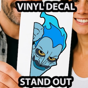 May include: A white vinyl decal featuring a cartoon illustration of Hades from Disney's Hercules. The character has blue skin and hair, with yellow eyes and a dark blue grin. The text "VINYL DECAL" and "STAND OUT" are displayed in black.