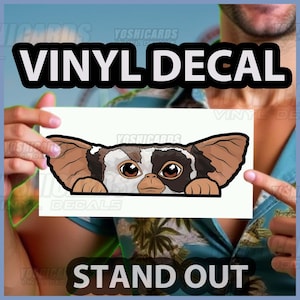 May include: A white vinyl decal featuring a Gremlin peeking over a ledge. The decal has brown, black, and white details. The words "VINYL DECAL" and "STAND OUT" are printed on the image.