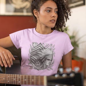 May include: A light pink t-shirt featuring a detailed black and white illustration of a Native American chief with a headdress, surrounded by floral elements. The design includes a star and striped pattern.