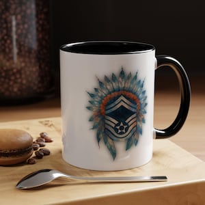 May include: White ceramic mug with a black interior and handle. The mug features a design of a blue and orange feathered headdress with a military insignia. A spoon and a pastry are on a wooden surface.