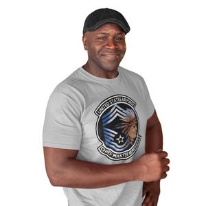 May include: A light gray t-shirt featuring a United States Air Force emblem with the text "Chief Master Sergeant." The emblem includes a Native American figure and blue and white stripes. The person in the image is wearing a black cap.