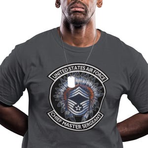 May include: A gray t-shirt featuring a United States Air Force emblem with the text "Chief Master Sergeant." The emblem includes a star and feather design. The person is wearing a silver chain.