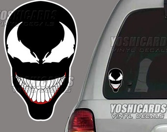 May include: Black and white vinyl decal featuring the Venom character's face. The design includes white eyes, a wide grin with white teeth, and a red outline for the mouth. The decal is shown on a car window.