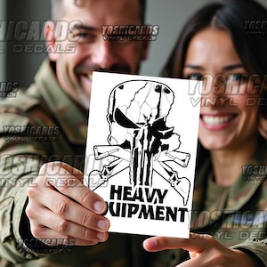 May include: A black and white vinyl decal featuring a skull design with crossed weapons and the words "HEAVY EQUIPMENT". The decal is held by two people in military-style attire.