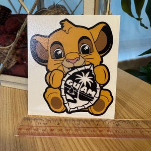 May include: A sticker featuring a cartoon lion cub holding a shield with the word "GUAM" and a palm tree design. The sticker is on a white background, with a ruler showing inches.
