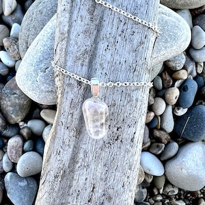 May include: A silver chain necklace with a clear quartz crystal pendant. The pendant is a natural stone with a light pink hue. The necklace is displayed on a piece of driftwood with small stones surrounding it.
