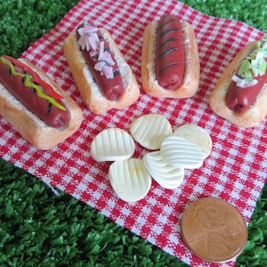 May include: Four miniature hot dogs on white bread rolls with various toppings, including onions, relish, and mustard, on a red and white checkered picnic blanket with a penny for scale.