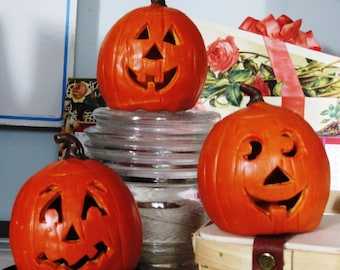 Può includere: Tre zucche di Halloween arancioni con volti intagliati e piccioli marroni. Una zucca è posizionata sopra una pila di vetro trasparente, mentre le altre due sono collocate su superfici inferiori. Ogni zucca presenta occhi triangolari e un sorriso.