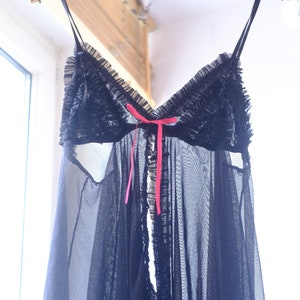 Könnte beinhalten: Schwarzes Dessous-Top mit Spaghettiträgern, Rüschenausschnitt und rosa Schleifenband. Der transparente Stoff ist mit zusätzlichen Rüschen am Saum versehen. Das Kleidungsstück hängt an einem Holzbügel.