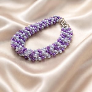 May include: A beaded bracelet with alternating rows of lavender and white beads. The bracelet is made of small, round beads and has a silver-colored clasp. The jewelry is displayed on a satin fabric.