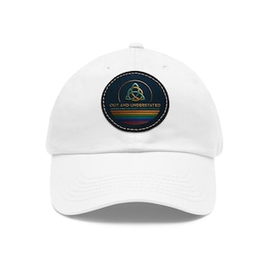 May include: White baseball cap with a circular patch featuring a gold and green Celtic knot design, the text "OUT AND UNDERSTATED", and a rainbow stripe design. The cap is made of a soft material and has a curved brim.