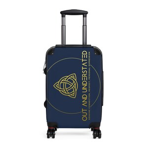 Peut inclure: Une valise bleu marine avec un cercle doré et un motif de nœud celtique doré. Le texte "OUT AND UNDERSTATED" est écrit verticalement en or sur le côté droit du cercle. Le texte "GENUINE CONNECTION, RESPECT, PERSONAL EXPRESSION" est écrit verticalement en or sur le côté droit de la valise.