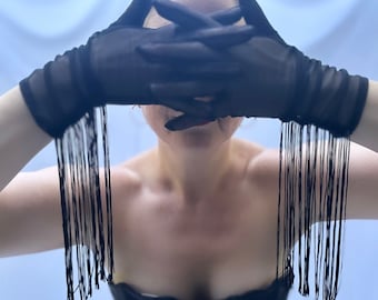 May include: A person wearing black fringed gloves covering their face. The gloves are made of a sheer fabric and have long, flowing fringes. The person is wearing a black strapless top.