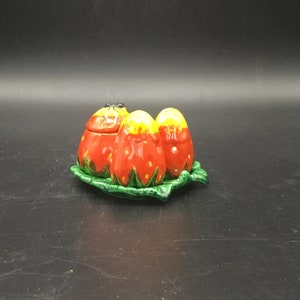 May include: A set of three ceramic strawberry-shaped salt and pepper shakers on a green leaf-shaped tray. The shakers are red and yellow with black eyes.