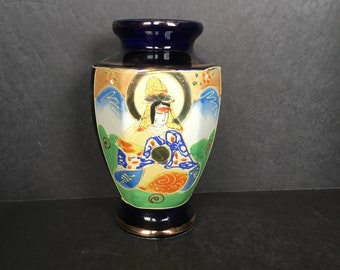 May include: A cobalt blue ceramic vase with a gold rim and a painted scene of a woman in a colorful robe. The vase has a hexagonal shape and is decorated with gold accents.
