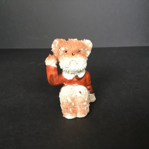May include: A small, brown and white ceramic figurine of a bear wearing a red jacket. The bear is sitting with its right paw raised.