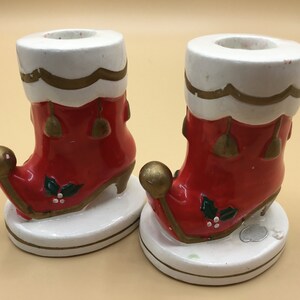 May include: Pair of red and white ceramic Christmas candle holders shaped like elf boots with gold trim and holly accents.