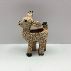 May include: A ceramic deer planter with a speckled brown and cream finish. The deer has large ears, a small tail, and black accents on its hooves and ears. The planter has a hollowed-out back for holding small plants or decorative items.