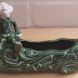 May include: A green ceramic planter shaped like a log with a small green and white ceramic gnome sitting on top. The gnome is smiling and has a pointed hat.