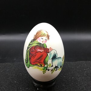 May include: A white, egg-shaped decorative item featuring a painted scene of a child in a red robe and green cape, interacting with a blue lamb. The artwork has a folk art style, with a black stand.