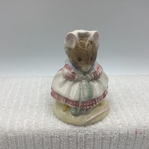 May include: A ceramic figurine of a mouse wearing a white dress with pink and red trim, a blue collar, and a white bonnet. The mouse is standing on a round, light-yellow base.