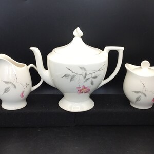 May include: A white teapot, creamer, and sugar bowl with a floral design. The design features a pink rose with gray leaves and stems.