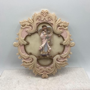 May include: A decorative wall plaque featuring a sculpted angel holding flowers and small animals. The angel is dressed in pink, blue, and white robes, set against a cream-colored background. The plaque has a detailed, ornate frame with pink and gold accents.