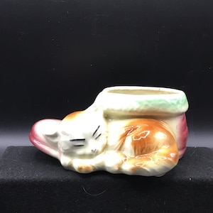 May include: A ceramic planter shaped like a sleeping cat. The cat is brown, white, and has green eyes. The planter has a green rim and a red base.