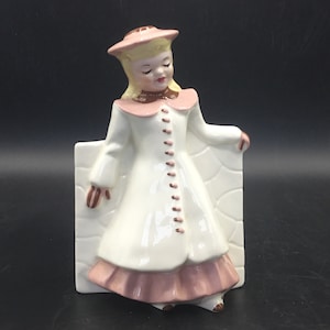 May include: A white ceramic wall planter shaped like a girl wearing a pink hat, white coat, and pink dress. The planter has brown button details and is leaning against a white brick wall.