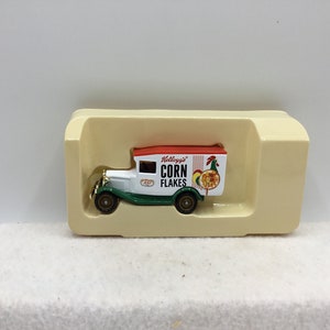 May include: A die-cast model of a Kellogg's Corn Flakes delivery truck. The truck is white with a red roof and green accents. The side of the truck features the Kellogg's logo and a rooster graphic. The model is in a beige display case.