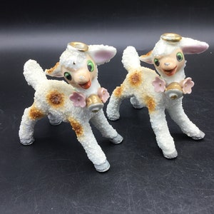 May include: Two white ceramic lamb figurines with brown spots, gold haloes, and pink flowers. Each lamb holds a gold bell in its mouth.