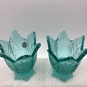 May include: Two teal-colored glass tulip-shaped bowls with detailed leaf patterns. Each bowl has a stem and a small label. The bowls are displayed on a white surface.