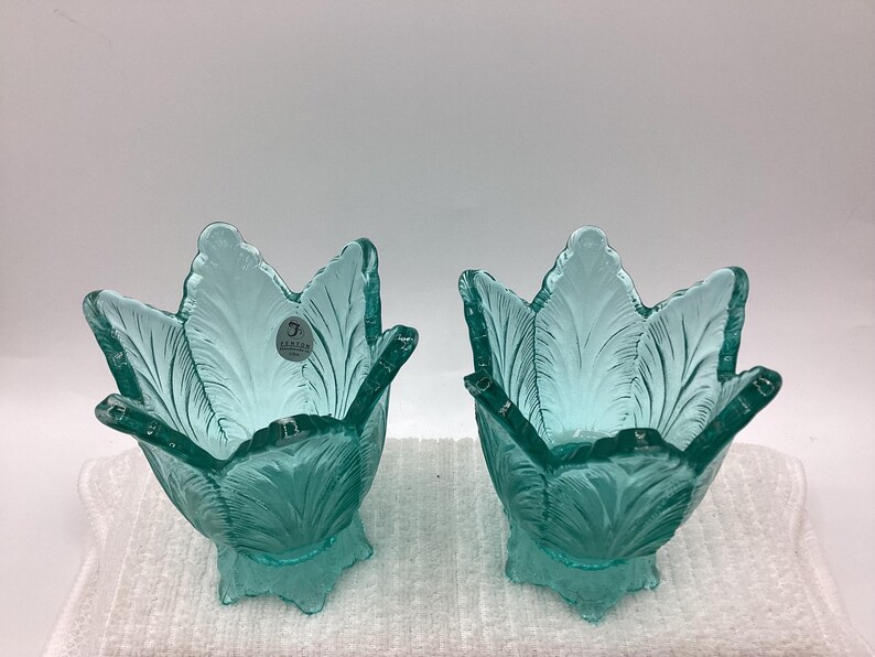 May include: Two teal-colored glass tulip-shaped bowls with detailed leaf patterns. Each bowl has a stem and a small label. The bowls are displayed on a white surface.