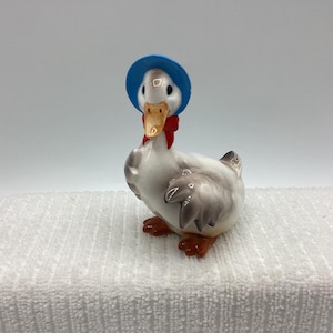 May include: A ceramic duck figurine wearing a blue bonnet and a red bow. The duck has white feathers, gray wings, and orange feet and beak. The figurine is on a white surface.