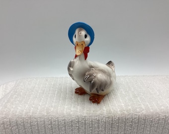May include: A ceramic duck figurine wearing a blue bonnet and a red bow. The duck has white feathers, gray wings, and orange feet and beak. The figurine is on a white surface.