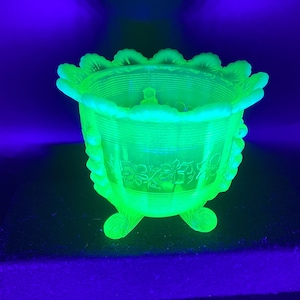 May include: A glowing green glass bowl with a scalloped rim and three small feet. The bowl has a floral design etched into the side and horizontal lines around the body. The bowl is illuminated with a bright green light.