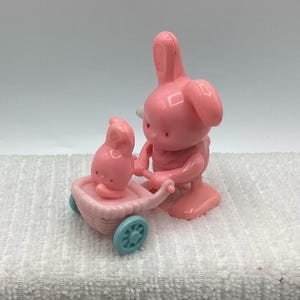 May include: A pink plastic bunny figure pushing a small pink stroller with a smaller bunny inside. The stroller has blue wheels. The bunny figure has large ears and is standing upright.