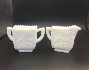 May include: A white milk glass sugar bowl and creamer set with a floral design. The pieces have a square shape with a raised rim and two handles.