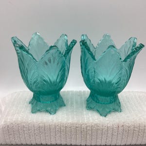 May include: Two turquoise glass tulip-shaped candle holders. The candle holders have a detailed leaf design and sit on a decorative base. The candle holders are displayed on a white towel.