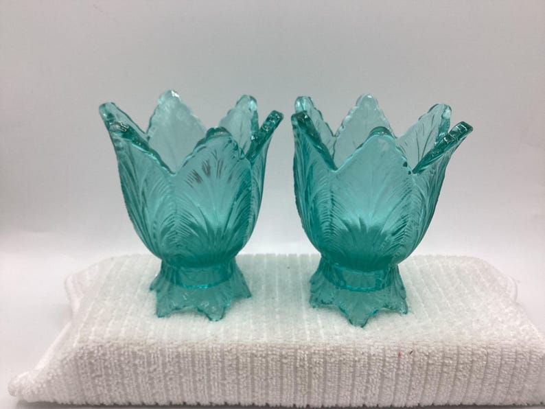 May include: Two turquoise glass tulip-shaped candle holders. The candle holders have a detailed leaf design and sit on a decorative base. The candle holders are displayed on a white towel.