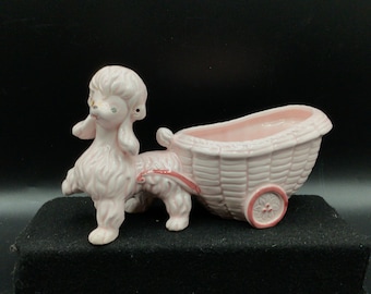 May include: A pink ceramic poodle figurine pulling a pink ceramic wagon planter. The wagon has a woven basket design and a single wheel.