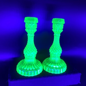 May include: Two matching green glass candlesticks with a textured, hobnail-like pattern. The candlesticks have a round base, a slender stem, and a top designed to hold a candle. The glass glows brightly against a dark blue background.