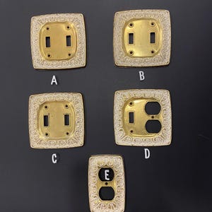 Puede incluir: Cinco placas de pared decorativas con marcos dorados ornamentados y fondos blancos. Las placas están etiquetadas con las letras A, B, C, D y E. Las placas A, B y C tienen dos interruptores de palanca. La placa D tiene un interruptor de palanca y una toma de corriente. La placa E tiene una toma de corriente.